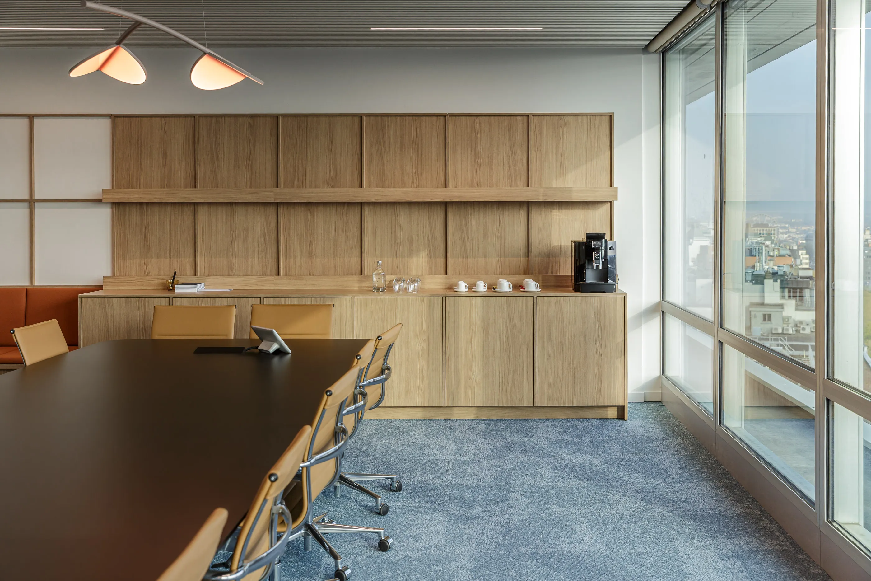 Salle de réunion ensoleillée avec table sombre, fauteuils fauves et rangements en bois sur toute la paroi