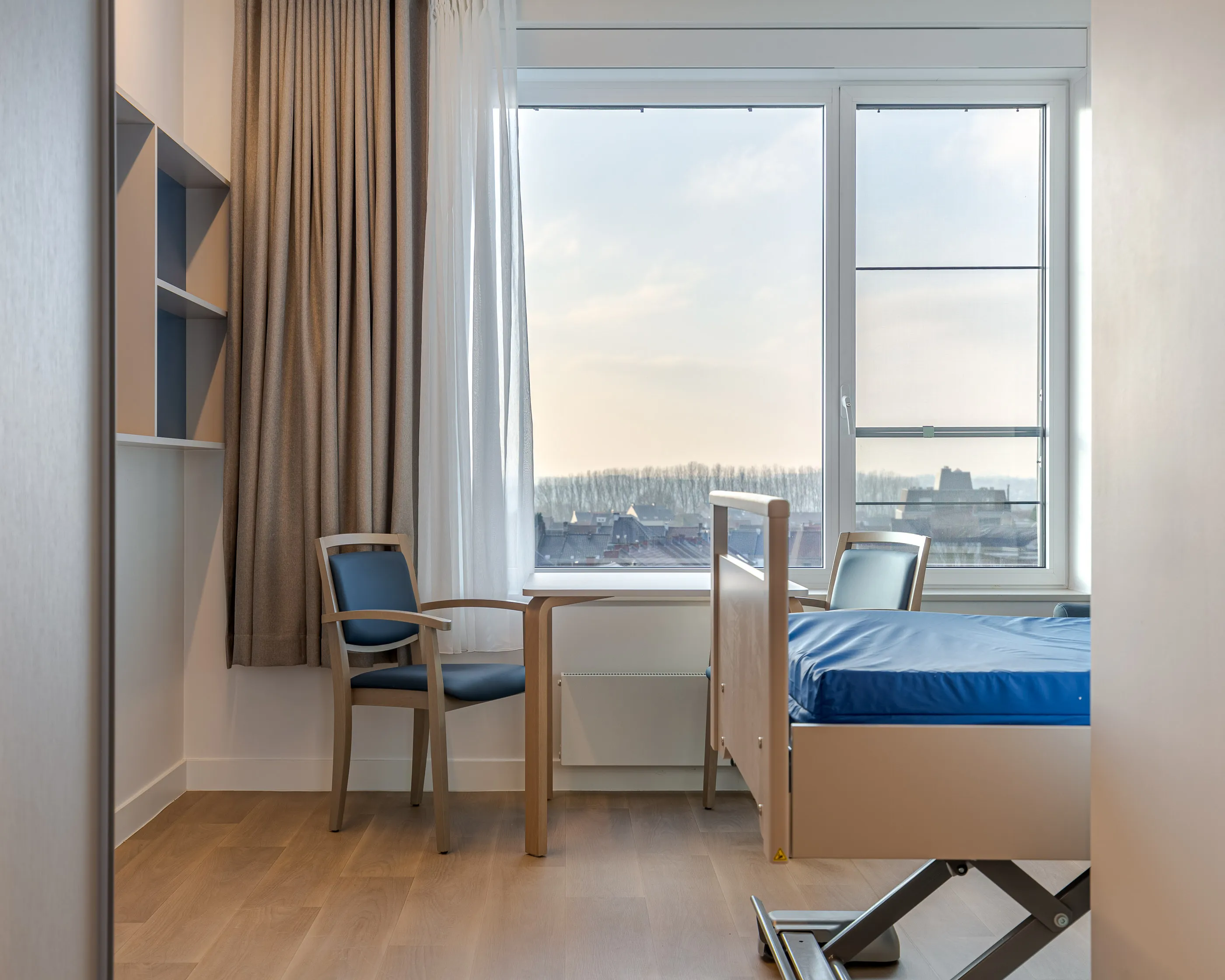 Chambre avec couvre-lit bleu, deux chaises et une table près d’une large fenêtre