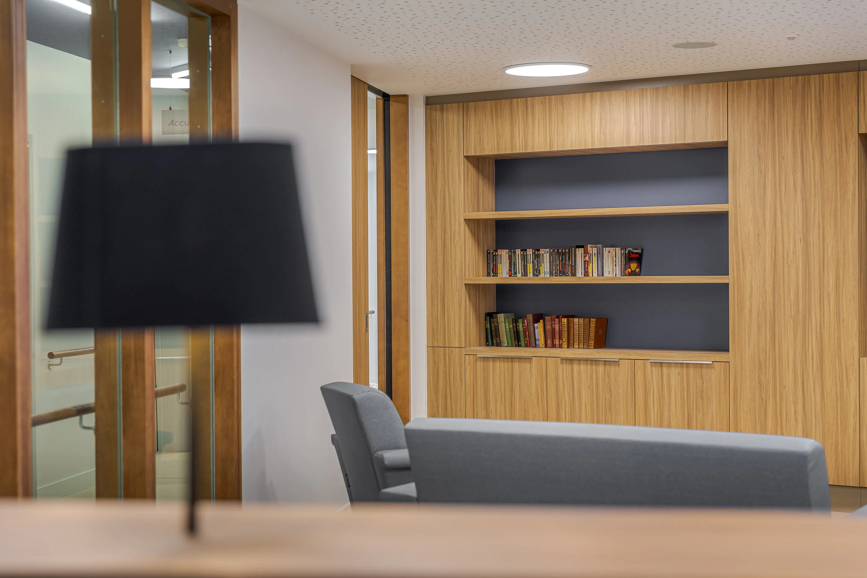 Salon avec sièges gris et bibliothèque en bois, cadré par les cloisons vitrées du couloir