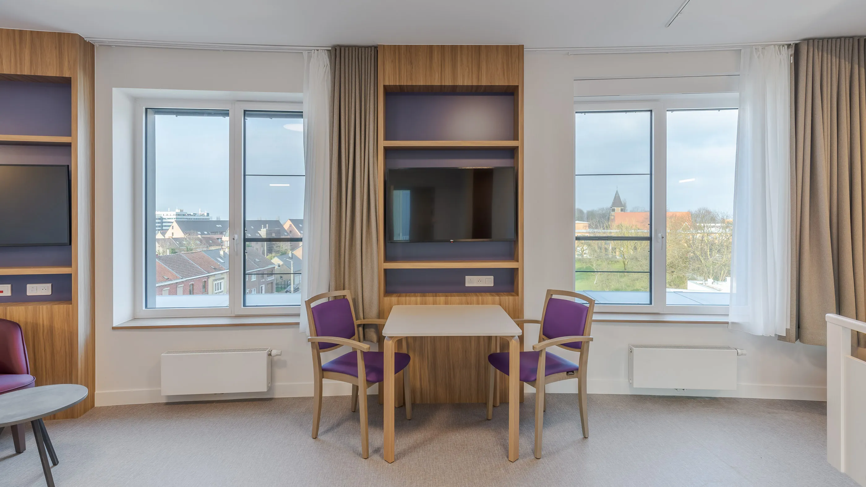 Deux fauteuils violets et une table entre de hautes fenêtres et des niches de télévision encadrées de bois
