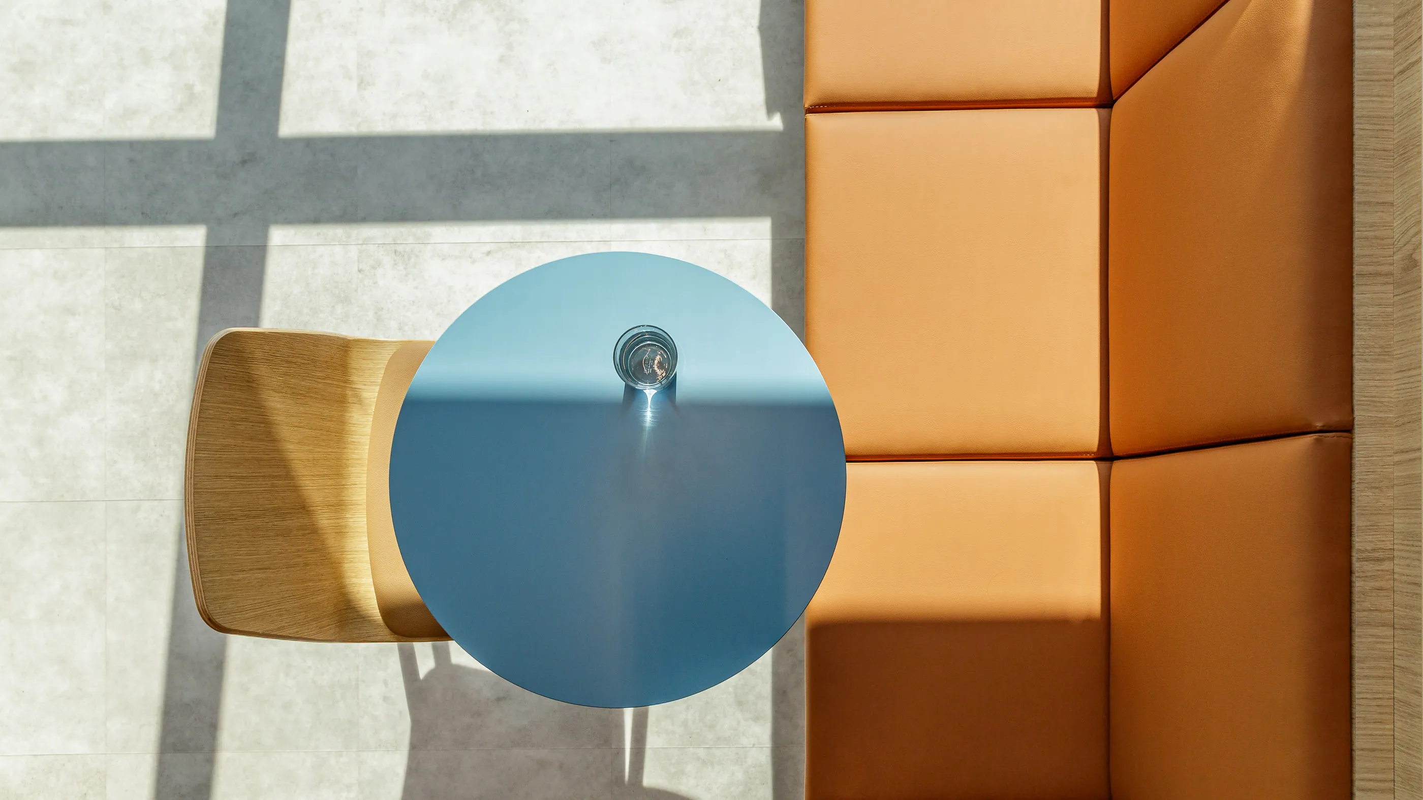 Vue plongeante sur une table ronde bleue et une banquette orange traversées par les ombres des fenêtres